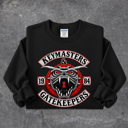 A black crewneck sweatshirt with a large graphic print on the front reading 'KEYMASTERS & GATEKEEPERS' in white and red, featuring a snarling horned face with a keyhole in the mouth. The sweatshirt is laid flat on a textured gray concrete surface.