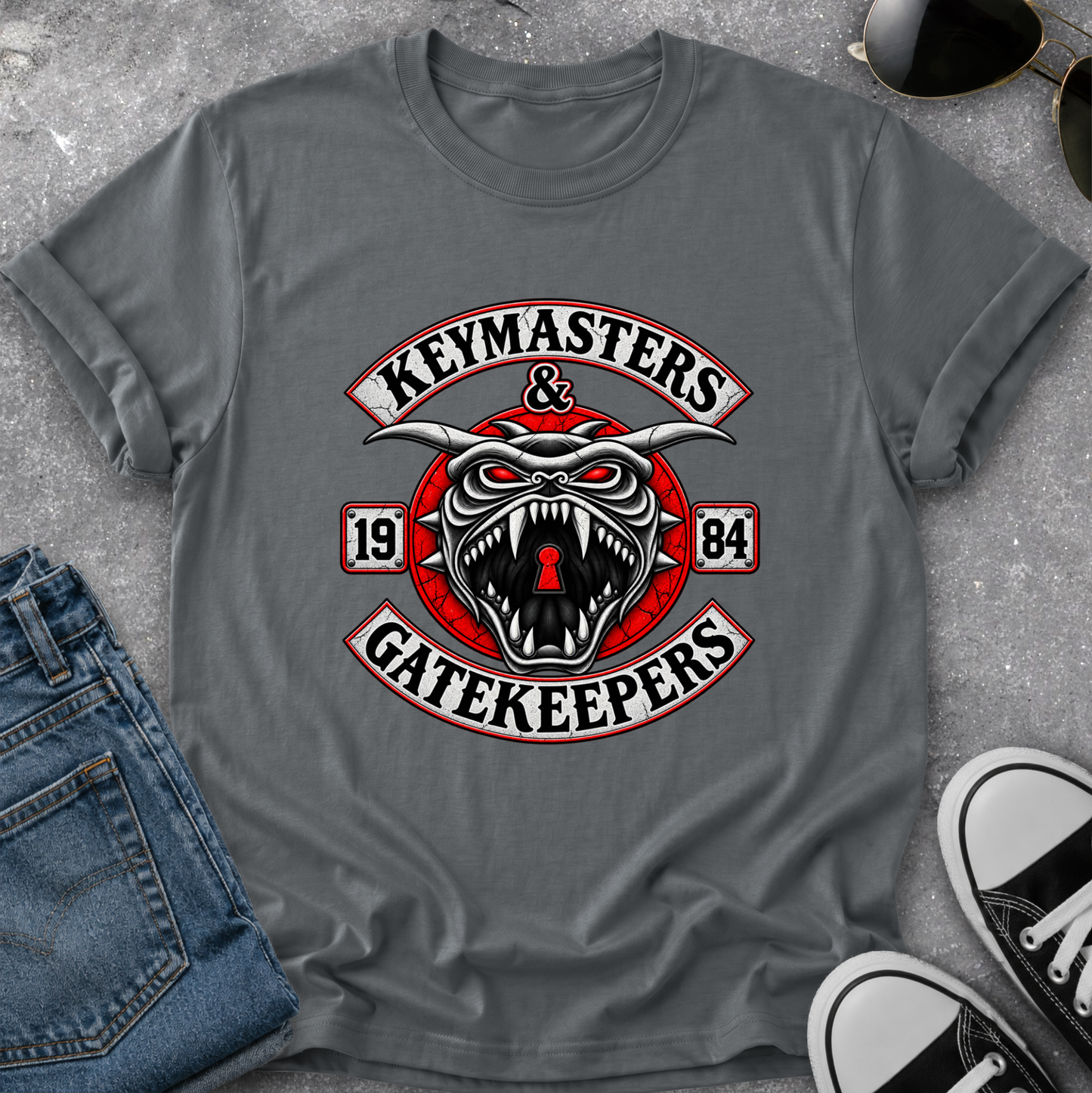 A gray short-sleeve t-shirt laid flat on a textured gray surface, featuring a large graphic print with a snarling horned skull, red circular elements, and the words 'KEYMASTERS & GATEKEEPERS' with '1984'. Blue jeans, sunglasses, and black-and-white sneakers are partially visible around it as styling props.