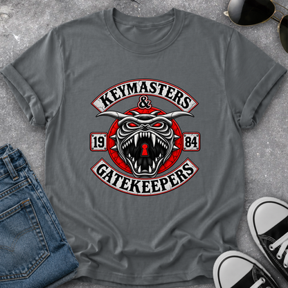 A gray short-sleeve t-shirt laid flat on a textured gray surface, featuring a large graphic print with a snarling horned skull, red circular elements, and the words 'KEYMASTERS & GATEKEEPERS' with '1984'. Blue jeans, sunglasses, and black-and-white sneakers are partially visible around it as styling props.