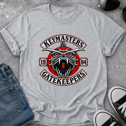 A heather gray short-sleeve t-shirt laid flat on a textured gray surface, featuring a large graphic print with a snarling horned creature, red circular elements, and the words 'Keymasters & Gatekeepers' with '1984'. Blue jeans, sunglasses, and black-and-white sneakers are partially visible around it as styling props.