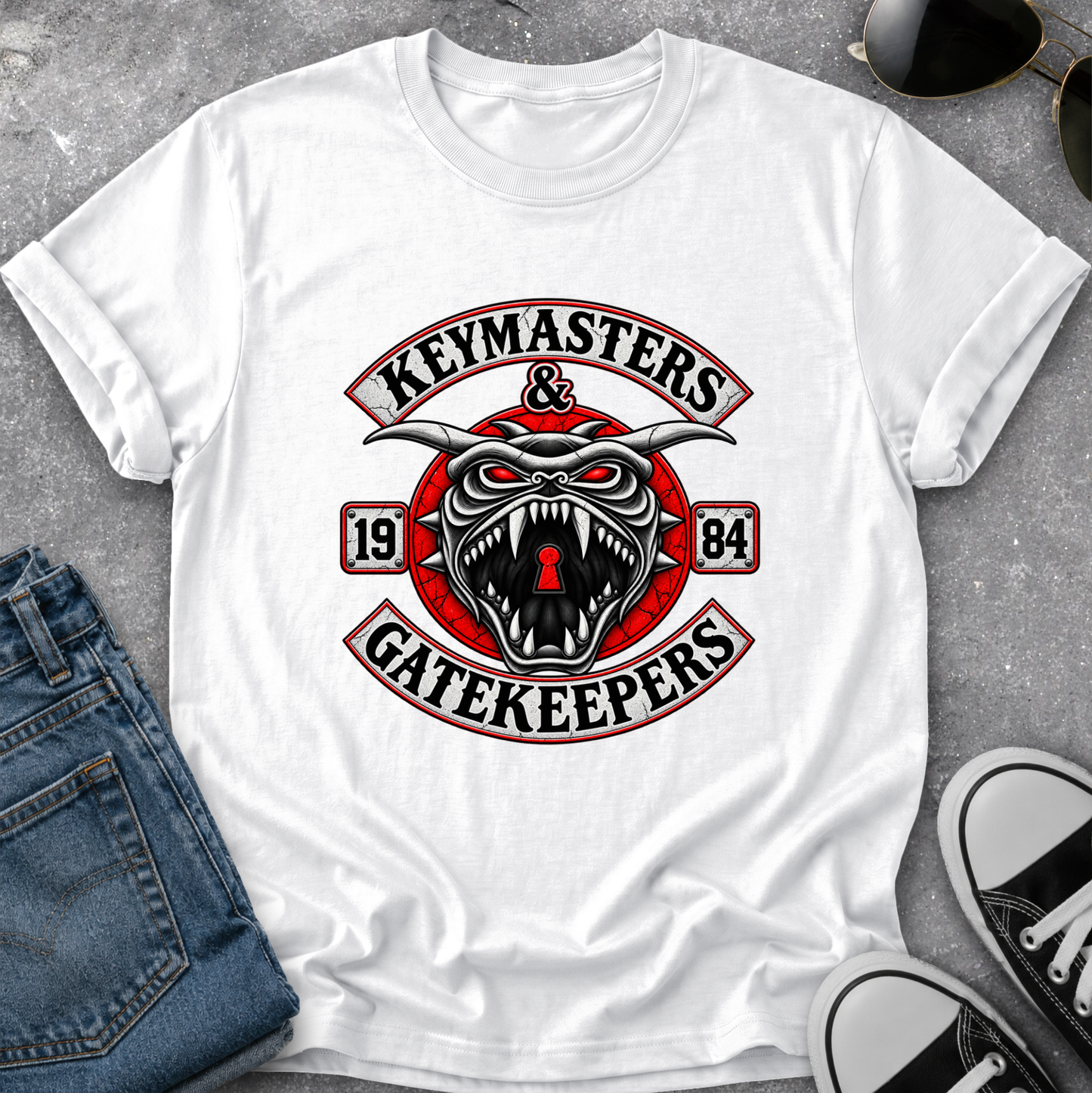 A white short-sleeve graphic T-shirt laid flat on a gray textured surface, styled with folded blue jeans, sunglasses, and black-and-white sneakers. The shirt features a large central graphic with the words 'KEYMASTERS & GATEKEEPERS' and a snarling keyhole-themed creature with red, black, and white details.