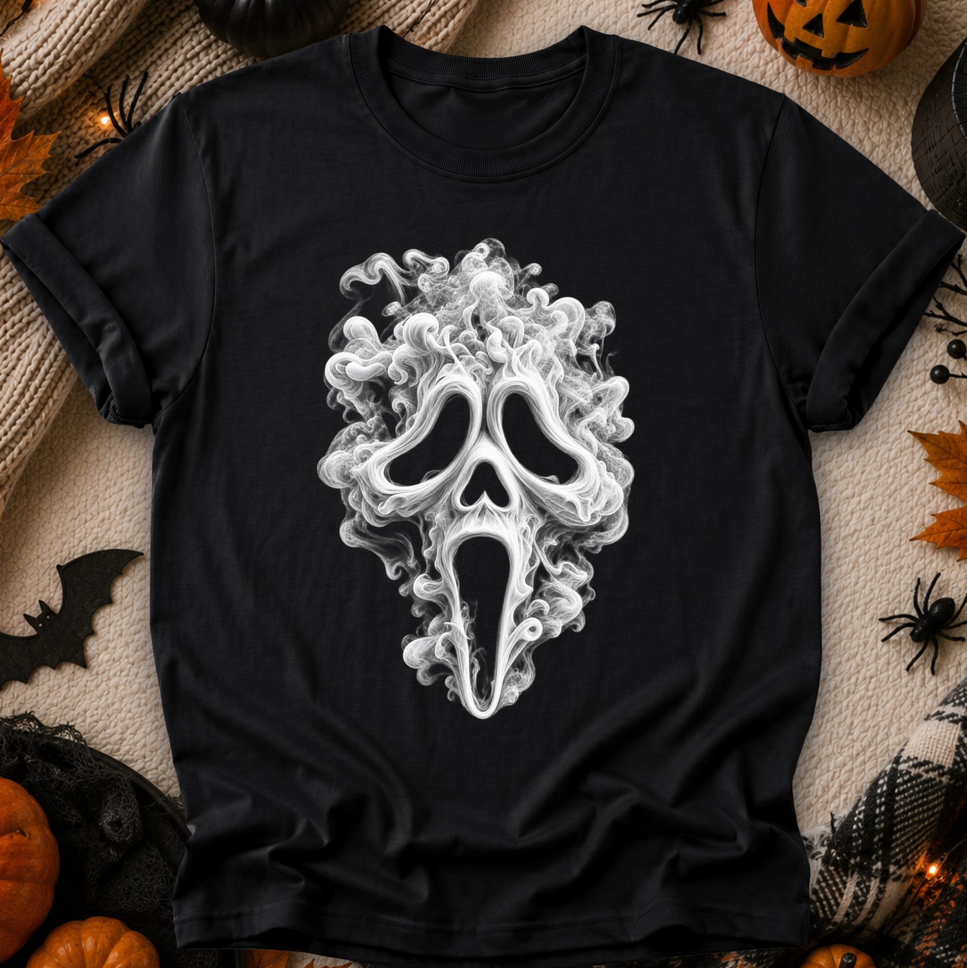 A black short-sleeve T-shirt laid flat with a large white Ghostface mask graphic centered on the front. The shirt is surrounded by Halloween-themed props including pumpkins, spider decorations, a black spiderweb, and warm string lights on a beige fabric surface.