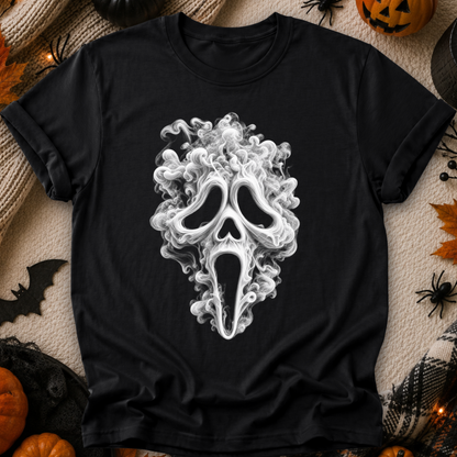 A black short-sleeve T-shirt laid flat with a large white Ghostface mask graphic centered on the front. The shirt is surrounded by Halloween-themed props including pumpkins, spider decorations, a black spiderweb, and warm string lights on a beige fabric surface.
