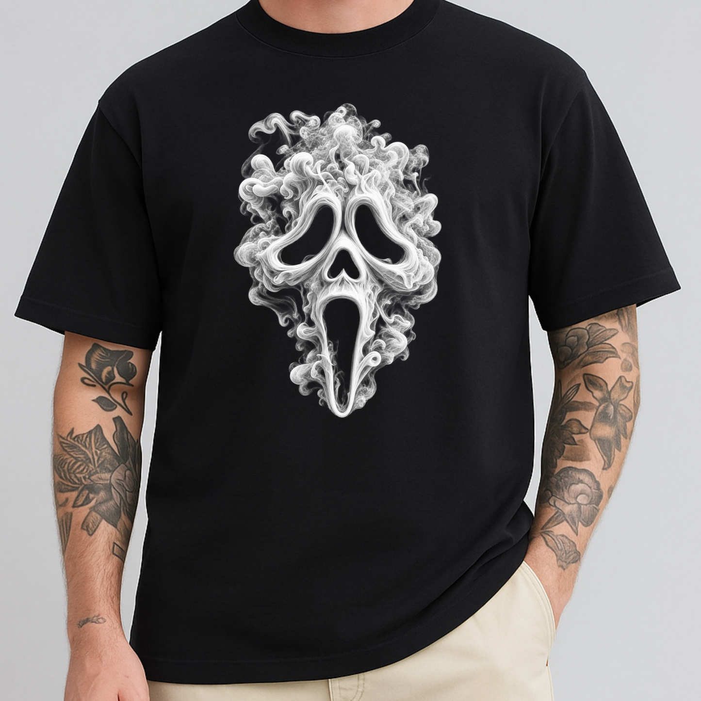 A man wearing a black short-sleeve t-shirt with a large white and gray smoke-spiral Ghostface mask graphic on the chest, paired with beige pants, against a solid gray background.