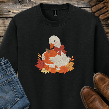 A black crewneck sweatshirt featuring a graphic of a white duck holding an orange pumpkin surrounded by autumn leaves and a red bow. The sweatshirt is styled in a flat-lay arrangement with blue jeans, brown leather shoes, and a pair of brown gloves on a rustic wooden background.