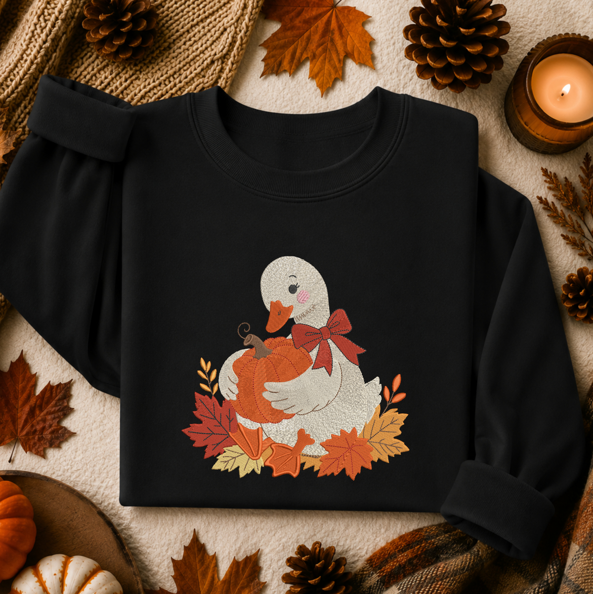 A black long-sleeve sweatshirt laid flat with rolled cuffs, featuring a central autumn-themed graphic of a white duck holding an orange pumpkin, surrounded by red and orange leaves and a red bow. The sweatshirt is styled in a cozy fall flat lay with pine cones, leaves, a candle, and small pumpkins.