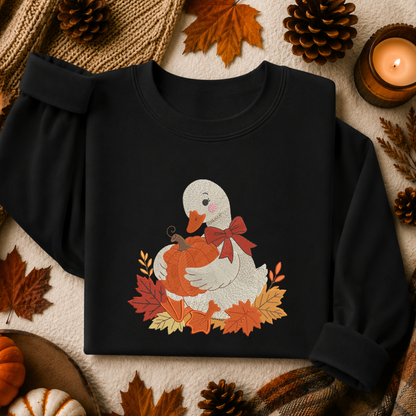 A black long-sleeve sweatshirt laid flat with rolled cuffs, featuring a central autumn-themed graphic of a white duck holding an orange pumpkin, surrounded by red and orange leaves and a red bow. The sweatshirt is styled in a cozy fall flat lay with pine cones, leaves, a candle, and small pumpkins.