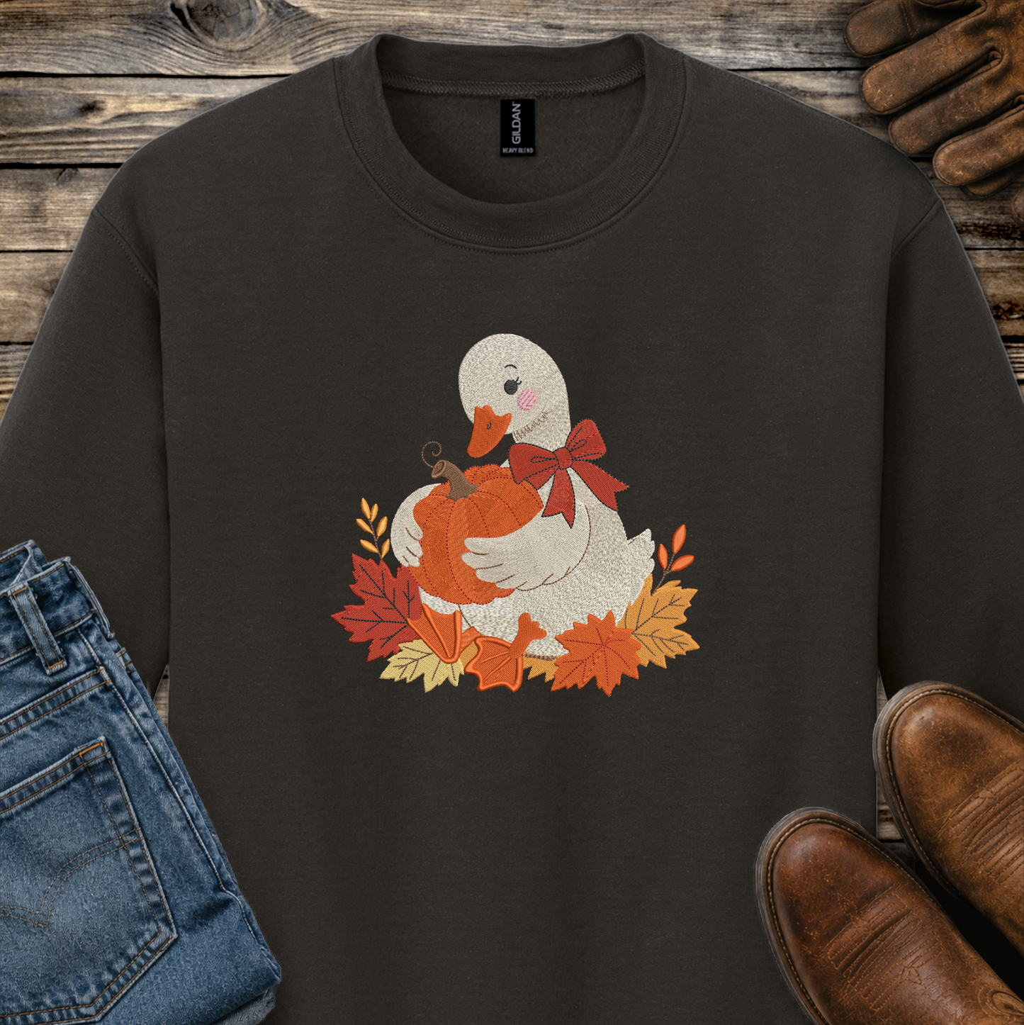 A dark brown crewneck sweatshirt featuring a graphic of a white goose holding an orange pumpkin surrounded by autumn leaves and a red bow. The sweatshirt is styled in a flat-lay arrangement with blue jeans, brown leather shoes, and a pair of brown gloves on a rustic wooden background.