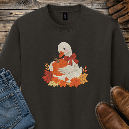 A dark brown crewneck sweatshirt featuring a graphic of a white goose holding an orange pumpkin surrounded by autumn leaves and a red bow. The sweatshirt is styled in a flat-lay arrangement with blue jeans, brown leather shoes, and a pair of brown gloves on a rustic wooden background.