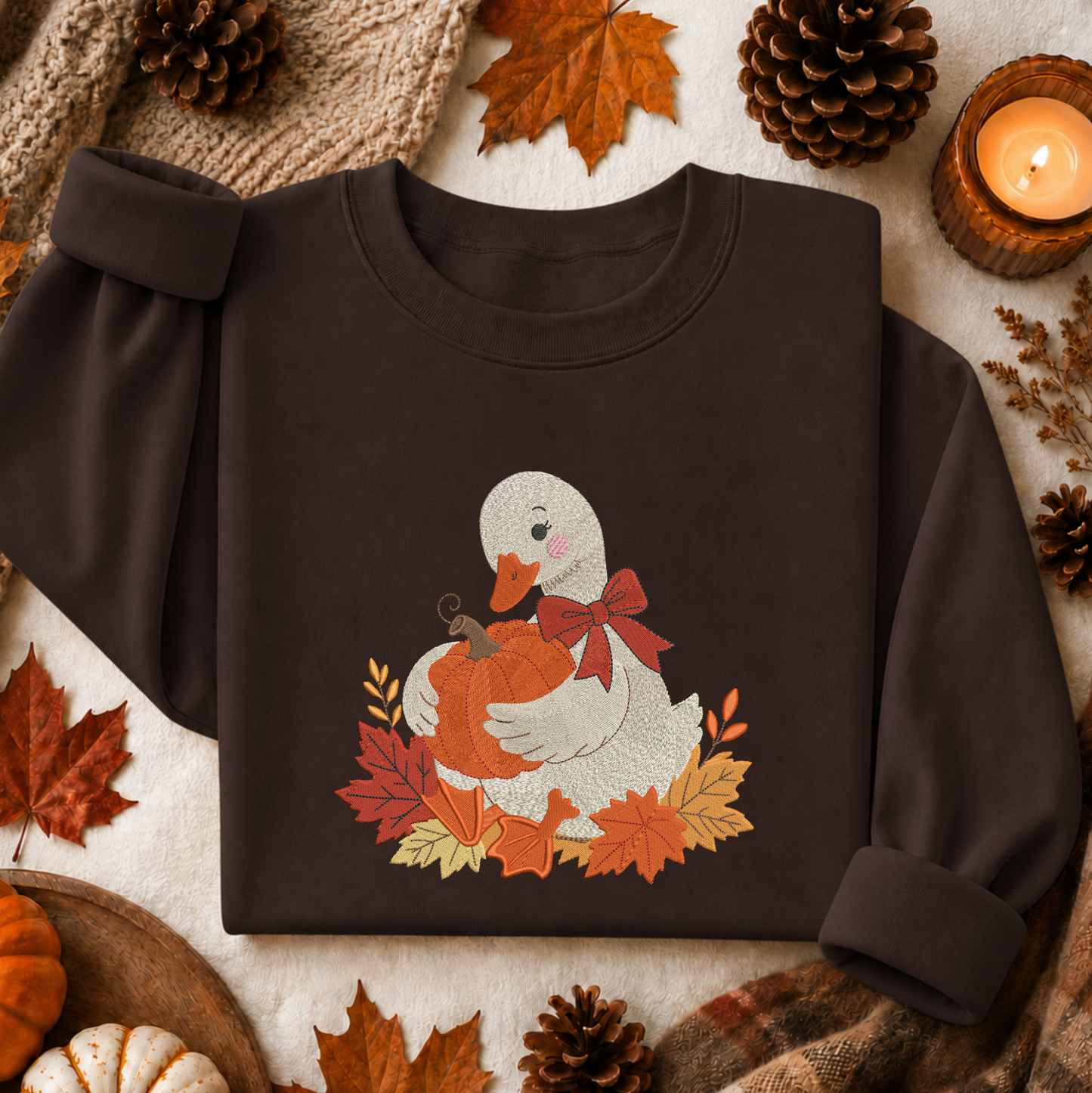 A dark brown crewneck sweatshirt laid flat on a neutral fabric background, decorated with autumn leaves, pinecones, a candle, and a knitted scarf. The sweatshirt features a graphic of a white duck holding an orange pumpkin, surrounded by red and orange autumn leaves and a red bow.