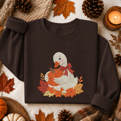 A dark brown crewneck sweatshirt laid flat on a neutral fabric background, decorated with autumn leaves, pinecones, a candle, and a knitted scarf. The sweatshirt features a graphic of a white duck holding an orange pumpkin, surrounded by red and orange autumn leaves and a red bow.