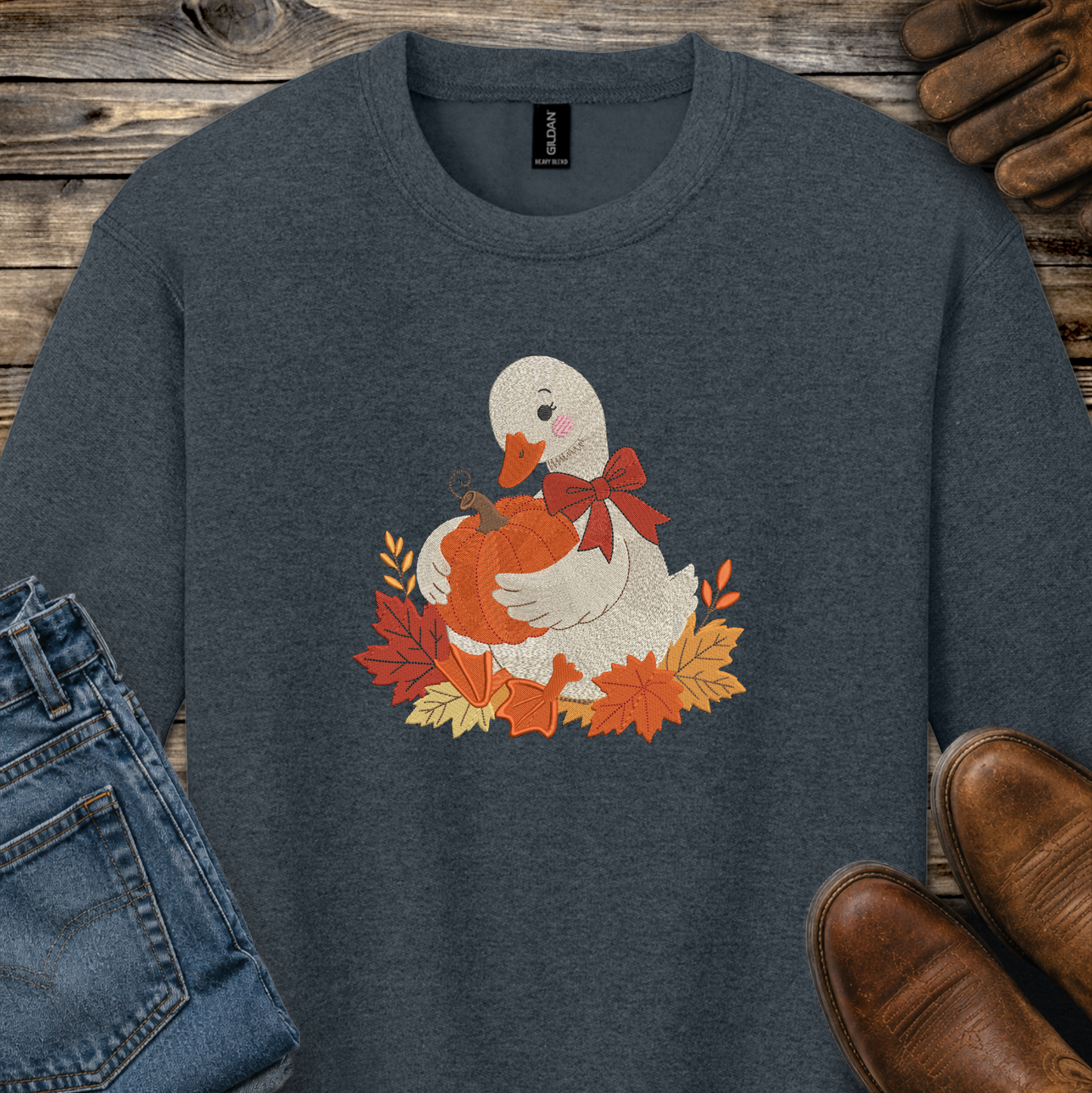 A dark heather gray crewneck sweatshirt featuring a graphic of a white duck holding an orange pumpkin surrounded by autumn leaves and a red bow. The sweatshirt is styled in a flat-lay arrangement with blue jeans, brown leather shoes, and a rustic wooden background.