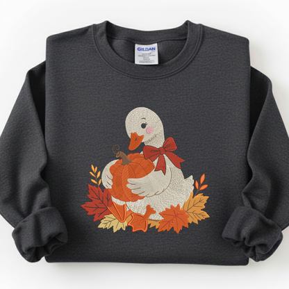 A dark charcoal gray crewneck sweatshirt featuring a graphic of a white duck holding an orange pumpkin surrounded by autumn leaves, with a red bow. The sweatshirt is laid flat on a white background, showing a Gildan brand label inside the collar.