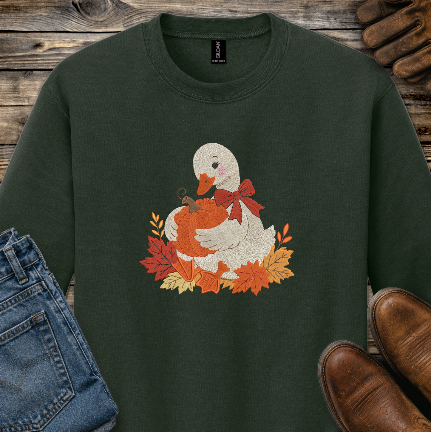 A dark forest green crewneck sweatshirt featuring a cute graphic of a white duck holding an orange pumpkin surrounded by autumn leaves and a red bow. The sweatshirt is styled in a flat-lay arrangement on a rustic wooden background, accompanied by blue jeans, brown leather shoes, and a pair of brown gloves.