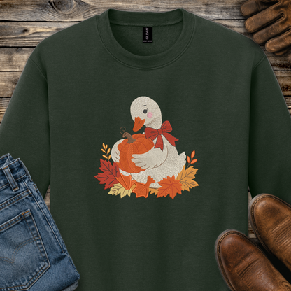 A dark forest green crewneck sweatshirt featuring a cute graphic of a white duck holding an orange pumpkin surrounded by autumn leaves and a red bow. The sweatshirt is styled in a flat-lay arrangement on a rustic wooden background, accompanied by blue jeans, brown leather shoes, and a pair of brown gloves.