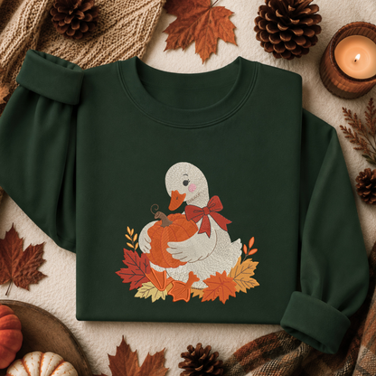 A dark forest green crewneck sweatshirt laid flat on a cozy autumn-themed background. The sweatshirt features a graphic of a white duck holding an orange pumpkin, surrounded by red and orange leaves and a red bow.