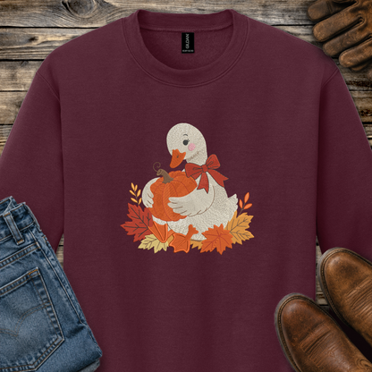 A maroon crewneck sweatshirt featuring a graphic of a white duck holding an orange pumpkin surrounded by autumn leaves and a red bow. The sweatshirt is styled in a flat-lay arrangement with blue jeans, brown leather shoes, and brown leather work gloves on a rustic wooden background.
