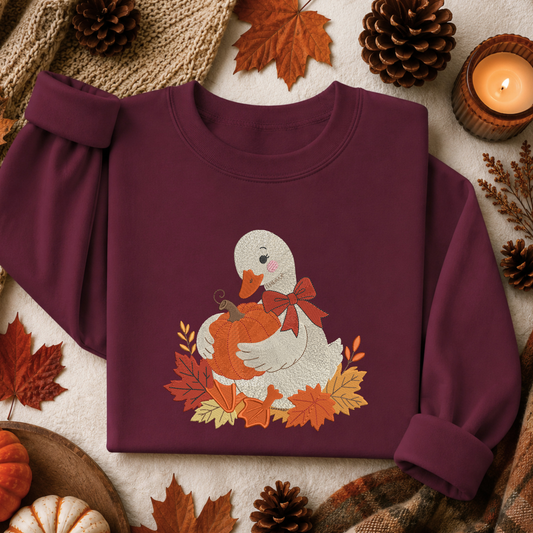A folded burgundy long-sleeve sweatshirt with a graphic of a white duck holding an orange pumpkin surrounded by autumn leaves and a red bow, styled on a neutral fabric background with pinecones, leaves, and a candle.