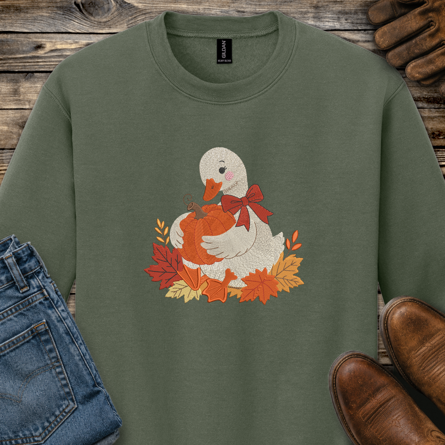 An olive green crewneck sweatshirt featuring a graphic of a white duck holding an orange pumpkin surrounded by autumn leaves and a red bow. The sweatshirt is styled in a flat-lay arrangement with blue jeans, brown shoes, and a wooden background.