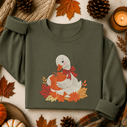 An olive green crewneck sweatshirt laid flat on a textured cream fabric background, surrounded by autumn props including pinecones, maple leaves, a small pumpkin, and a lit candle. The sweatshirt features a graphic of a white duck holding an orange pumpkin, decorated with a red bow and surrounded by colorful fall leaves.