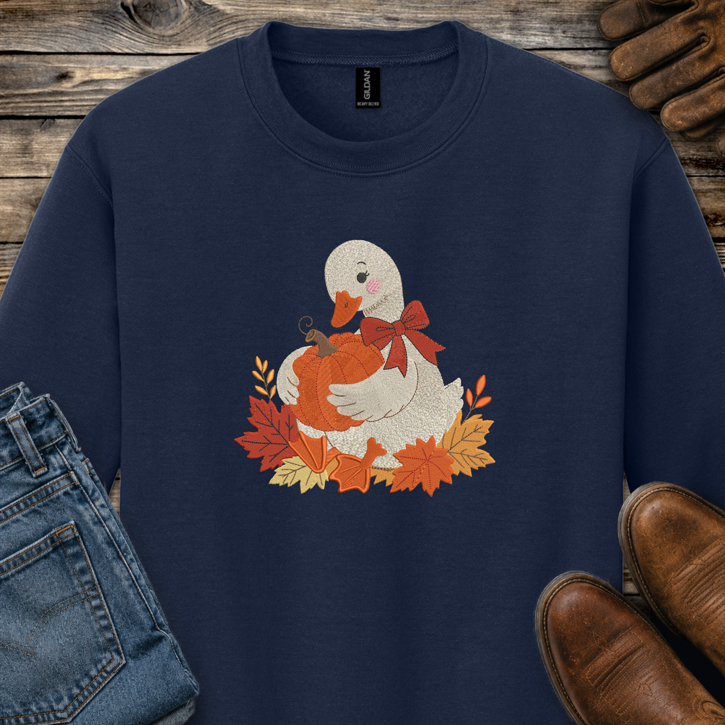 A navy blue crewneck sweatshirt featuring a graphic of a white duck holding an orange pumpkin surrounded by autumn leaves and a red bow. The sweatshirt is styled in a flat-lay arrangement on a rustic wooden background with blue jeans, brown leather shoes, and a pair of brown gloves.