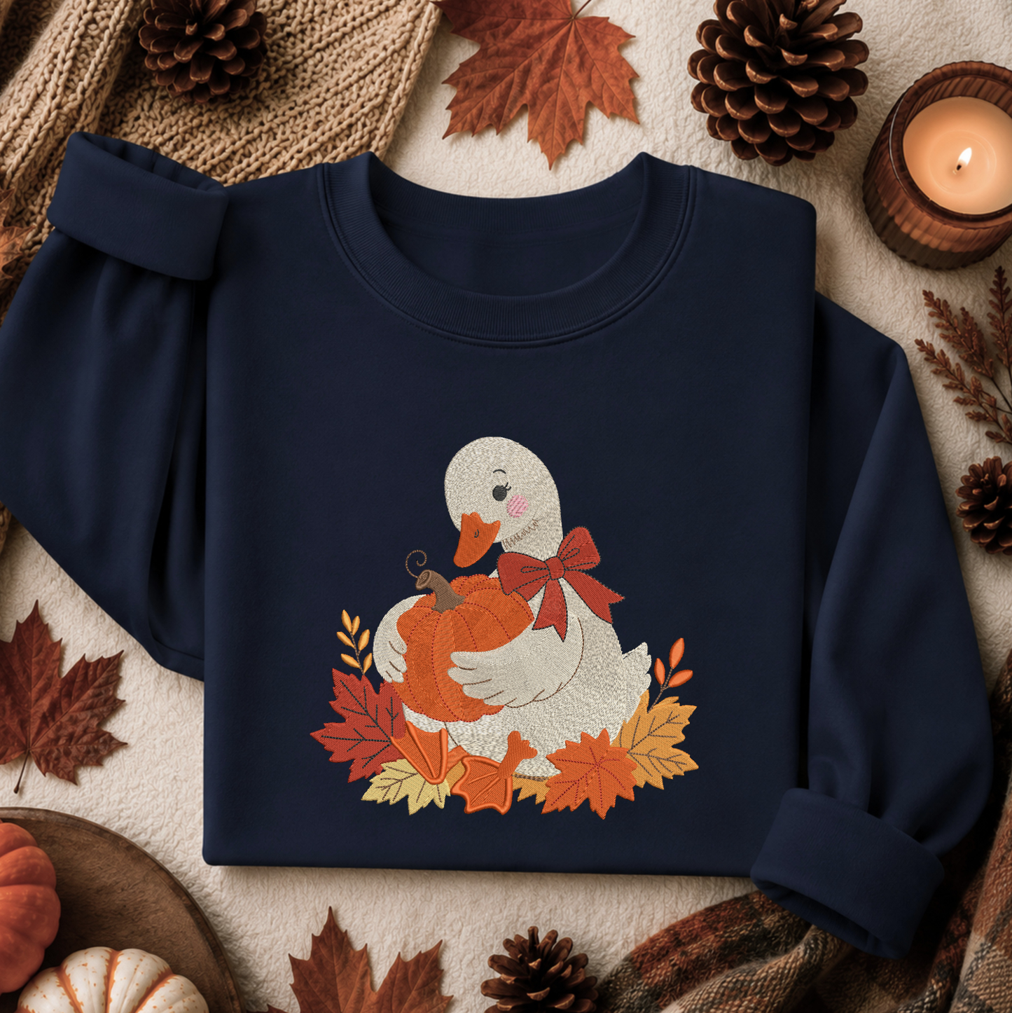 A navy blue long-sleeve sweatshirt laid flat on a cozy autumn-themed background. The sweatshirt features a graphic of a white duck holding an orange pumpkin, surrounded by orange and yellow leaves and a red bow.