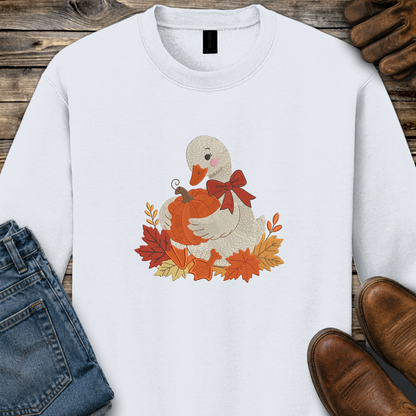 A white crewneck sweatshirt laid flat on a rustic wood surface, featuring a central graphic of a white duck holding an orange pumpkin surrounded by red, orange, and yellow autumn leaves and a red bow. Styled flat-lay props include blue jeans, brown shoes, and brown gloves.