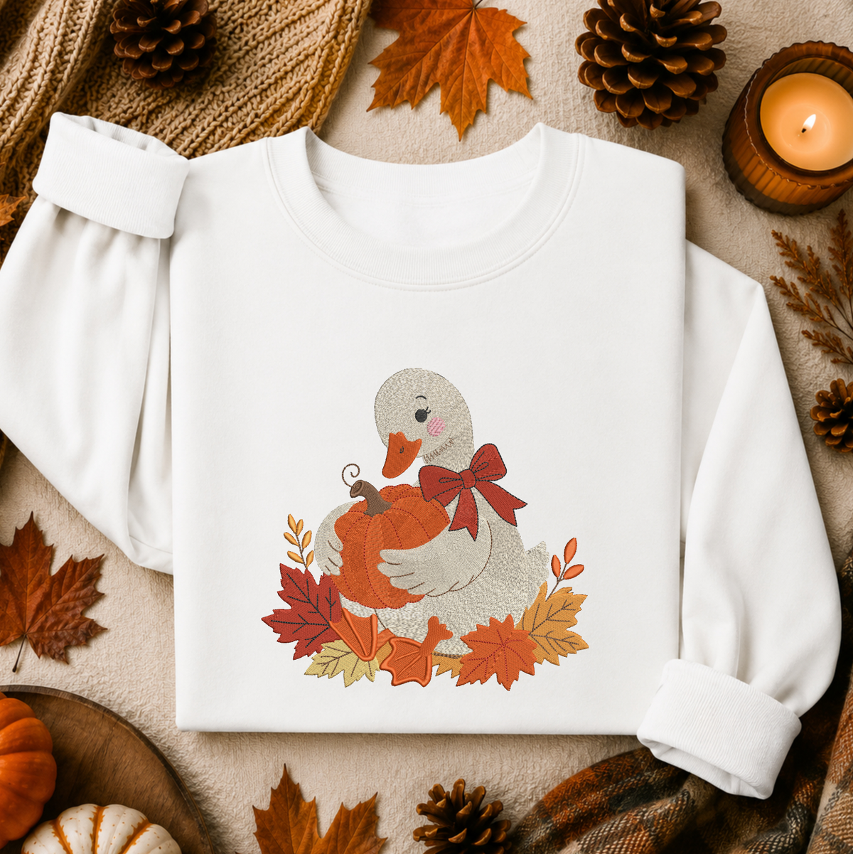 A white long-sleeve crewneck sweatshirt laid flat on a cozy autumn-themed background. The front features a graphic of a gray duck holding an orange pumpkin, surrounded by red and orange leaves and a red bow.