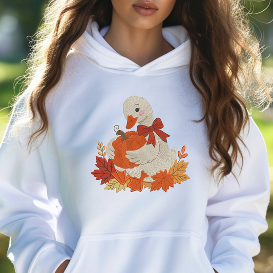A woman wearing a white pullover hoodie with a colorful autumn graphic of a duck holding an orange pumpkin surrounded by red, orange, and yellow leaves. The photo is taken outdoors with a soft, blurred green background.