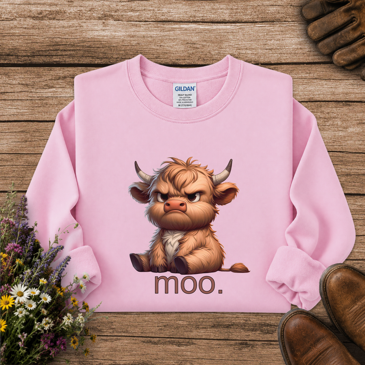 A pink long-sleeve sweatshirt laid flat on a rustic wooden surface, featuring a cartoon Highland cow graphic with the word 'moo.' underneath.