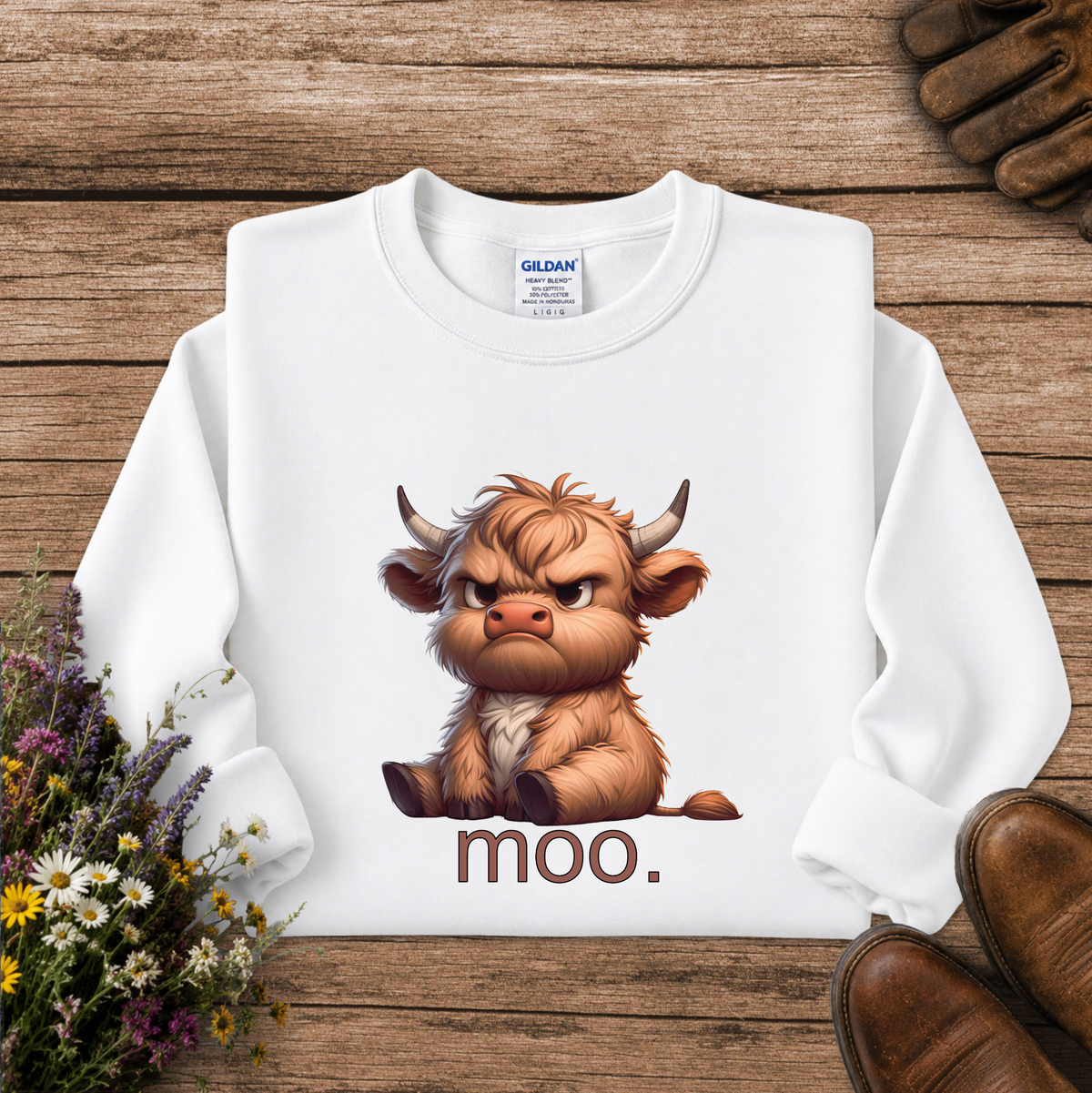 A white long-sleeve sweatshirt laid flat on a rustic wooden surface, featuring a cartoon illustration of an angry brown Highland cow with the word 'moo.' printed below.