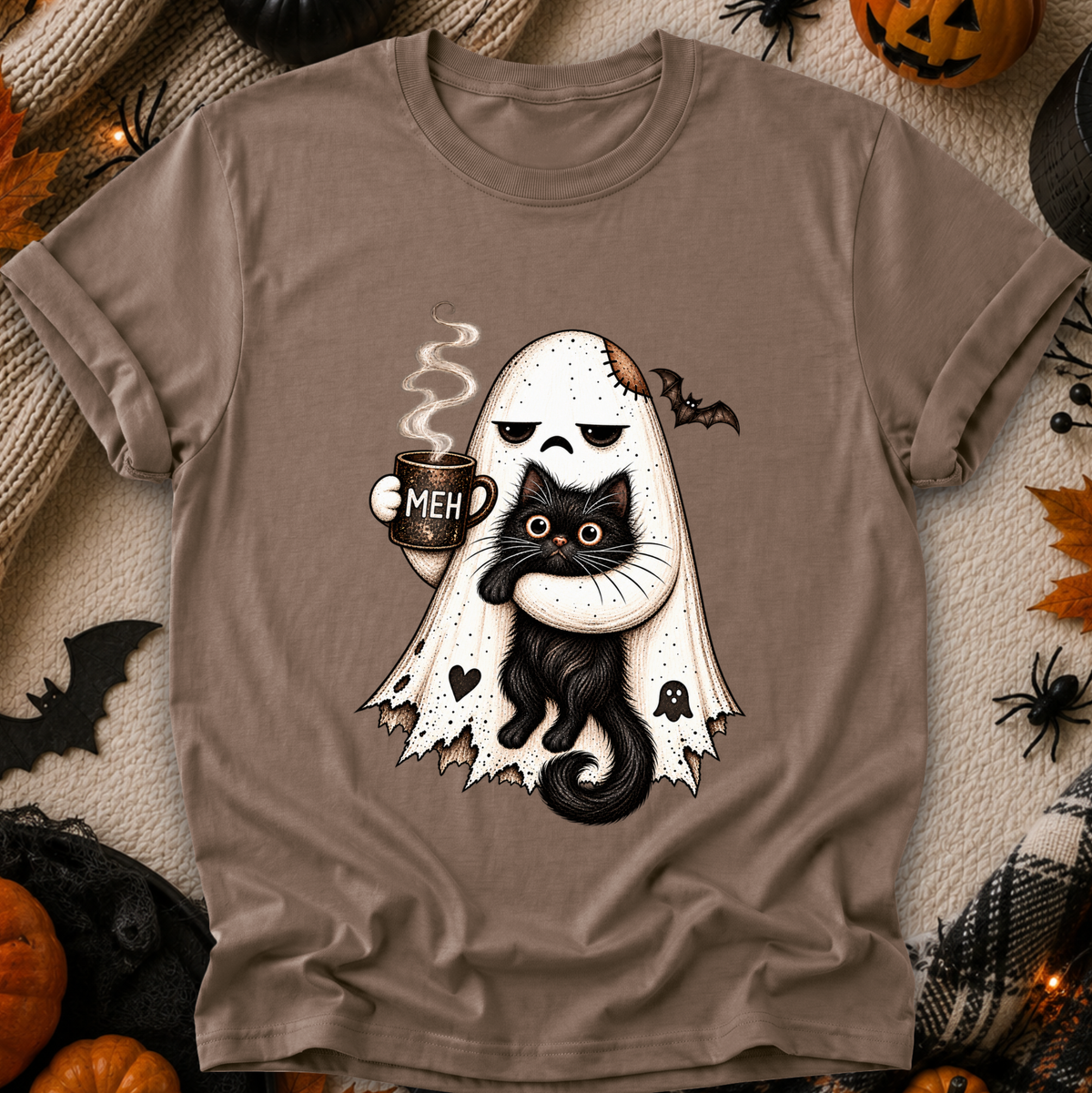 A taupe brown short-sleeve t-shirt with a Halloween graphic of a ghost hugging a black cat while holding a mug that says 'MEH'. The shirt is displayed in a flat-lay arrangement with autumn and Halloween props including pumpkins, spider decorations, and a knit blanket.