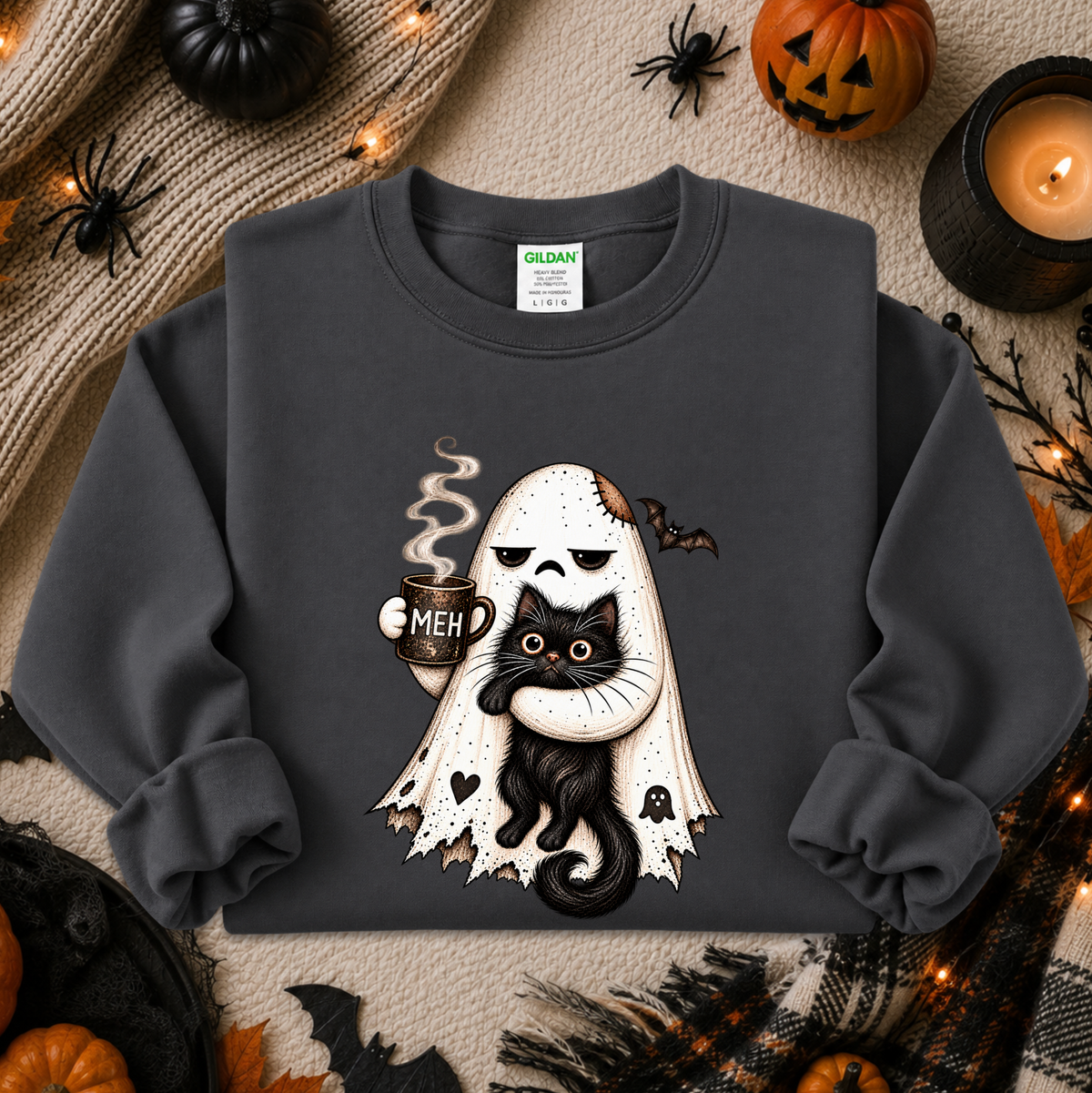 A dark charcoal gray crewneck sweatshirt featuring a Halloween graphic of a white ghost with tattered edges holding a black cat with glowing yellow eyes and a mug that says 'MEH'. The sweatshirt is displayed in a flat-lay arrangement surrounded by seasonal props including pumpkins, a candle, spider decorations, and a cozy knit blanket.