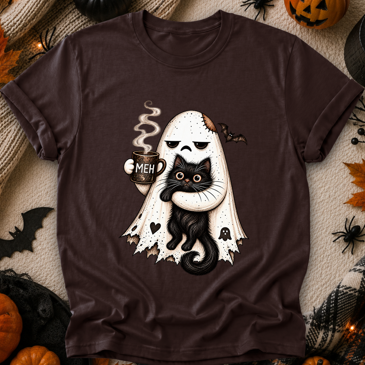 A dark brown short-sleeve t-shirt with a Halloween graphic of a ghost hugging a black cat while holding a mug that says 'MEH'. The shirt is displayed in a flat-lay arrangement with seasonal props like pumpkins, spider decorations, and a plaid blanket.