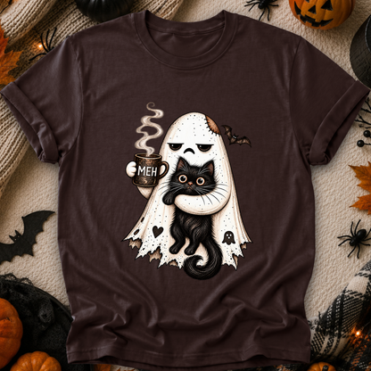 A dark brown short-sleeve t-shirt with a Halloween graphic of a ghost hugging a black cat while holding a mug that says 'MEH'. The shirt is displayed in a flat-lay arrangement with seasonal props like pumpkins, spider decorations, and a plaid blanket.