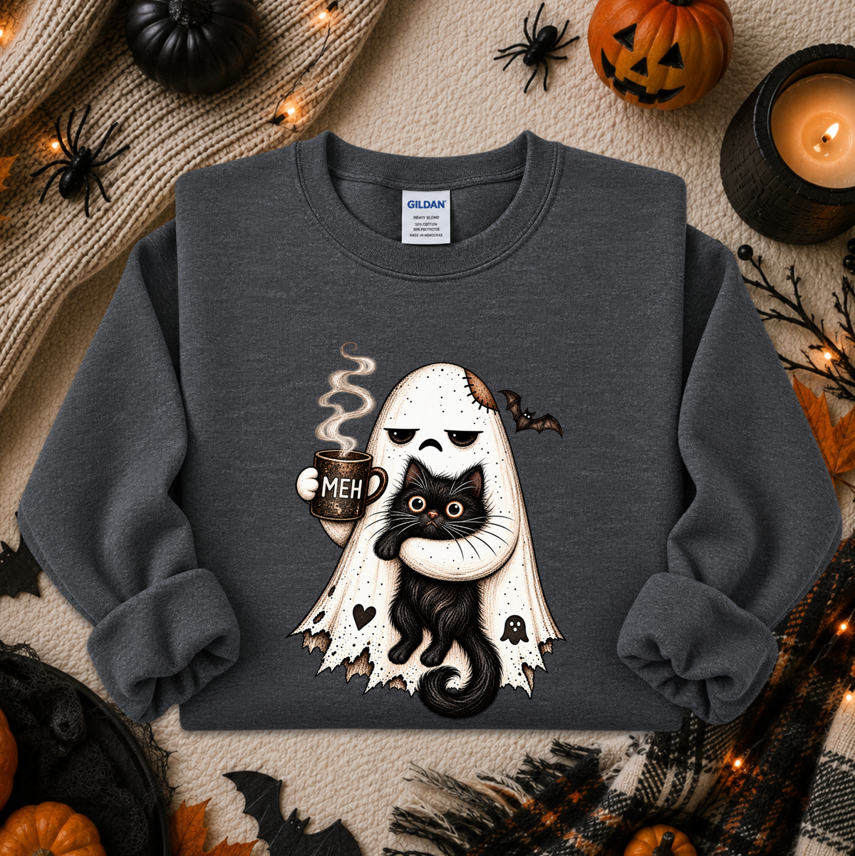 A dark gray crewneck sweatshirt featuring a cute graphic of a white ghost with a skull face holding a black cat and a mug that says 'MEH'. The sweatshirt is presented in a flat-lay Halloween setting with pumpkins, spider decorations, and a candle.
