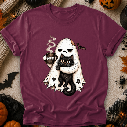 A mauve-purple short-sleeve t-shirt with a Halloween graphic of a ghost hugging a black cat while holding a mug that says 'MEH'. The shirt is displayed flat on a cozy beige blanket surrounded by autumn and Halloween props like pumpkins, spiderwebs, and a black spider decoration.