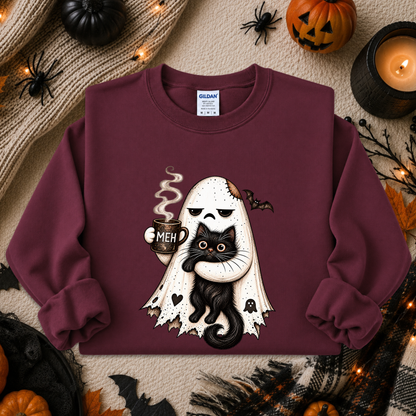 A maroon crewneck sweatshirt featuring a cartoon graphic of a white ghost holding a black cat with the text 'MEH', styled in a flat lay with Halloween props like pumpkins, a candle, and spider decorations. The brand Gildan is visible on the inner neck label.