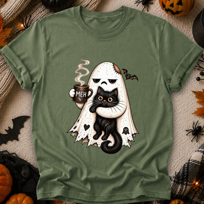 An olive green short-sleeve t-shirt with a cute Halloween graphic of a ghost hugging a black cat while holding a mug that says 'MEH'. The t-shirt is displayed in a flat-lay arrangement surrounded by seasonal props like pumpkins, spiderwebs, and a knitted blanket.