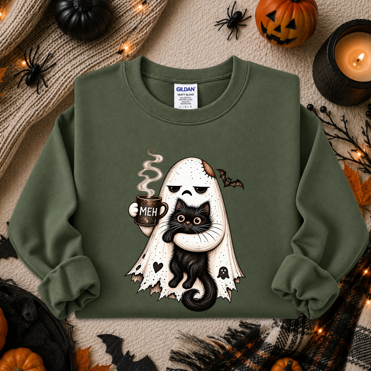 An olive green crewneck sweatshirt featuring a cartoon graphic of a ghost holding a black cat with a 'MEH' mug, styled in a flat lay with Halloween props like pumpkins, a candle, and spider decorations. The Gildan brand label is visible inside the collar.