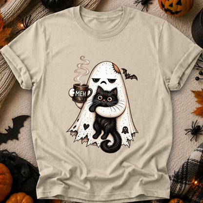A beige graphic t-shirt laid flat on a cozy, Halloween-themed background. The shirt features a cute cartoon illustration of a ghost hugging a black cat, with a mug that says 'MEH' and small bats.