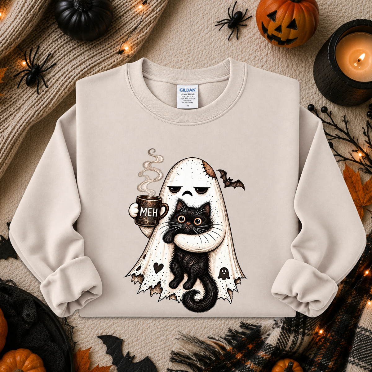 A cream-colored crewneck sweatshirt featuring a graphic of a white ghost with tattered edges holding a black cat and a mug that says 'MEH'. The sweatshirt is displayed in a flat-lay arrangement with Halloween-themed props including pumpkins, a candle, spider decorations, and autumn leaves.