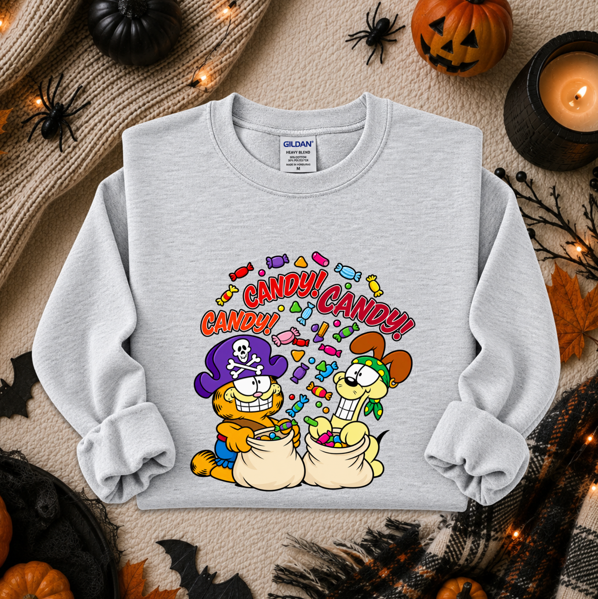 A light gray crewneck sweatshirt featuring a colorful cartoon graphic of Garfield and Catbus Man eating candy, styled in a cozy flat-lay with Halloween decorations.