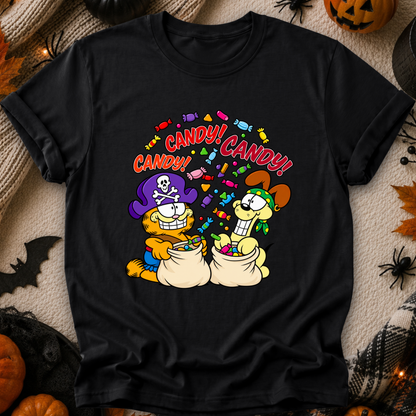 A black short-sleeve graphic T-shirt laid flat on a Halloween-themed background with pumpkins, spiders, and autumn leaves. The shirt features a colorful cartoon print of two cats sitting in bags of candy under the word 'CANDY!' repeated, with one cat wearing a purple pirate hat and the other wearing a brown witch hat.