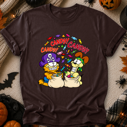 A dark brown short-sleeve graphic t-shirt featuring Garfield and Odie dressed as Halloween pirates with the text 'CANDY! CANDY!' above them. The shirt is displayed in a flat-lay arrangement on a cozy beige blanket surrounded by seasonal Halloween props like pumpkins, a spider, and a bat.