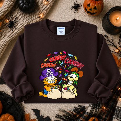 A dark brown crewneck sweatshirt featuring a colorful cartoon graphic of Garfield and Odie as trick-or-treaters with the text 'CANDY! CANDY!'. The sweatshirt is displayed in a flat-lay Halloween setting with pumpkins, a candle, and spider decorations.