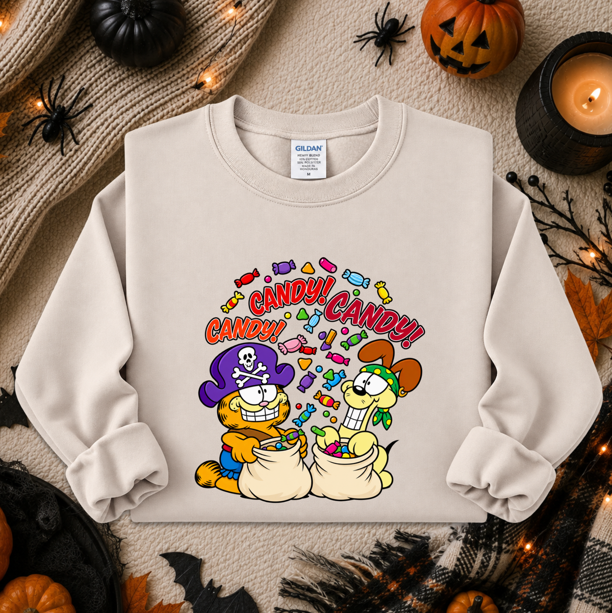 A cream-colored crewneck sweatshirt featuring a colorful cartoon graphic of Garfield and Catbus Charlie as pirates collecting candy, surrounded by Halloween props like pumpkins, a candle, and spider decorations.