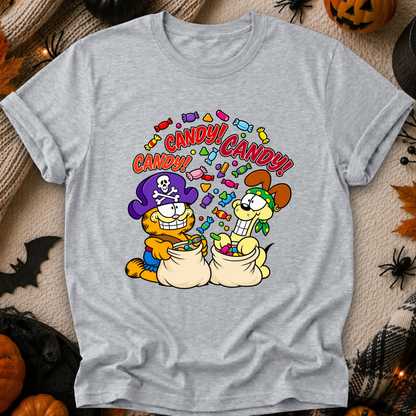 A light heather gray short-sleeve t-shirt with a colorful cartoon graphic of two dogs dressed as pirates and cats, surrounded by candy and the text 'CANDY! CANDY!'. The shirt is displayed flat on a cozy beige fabric background decorated with Halloween props like pumpkins, a spider, and a bat.