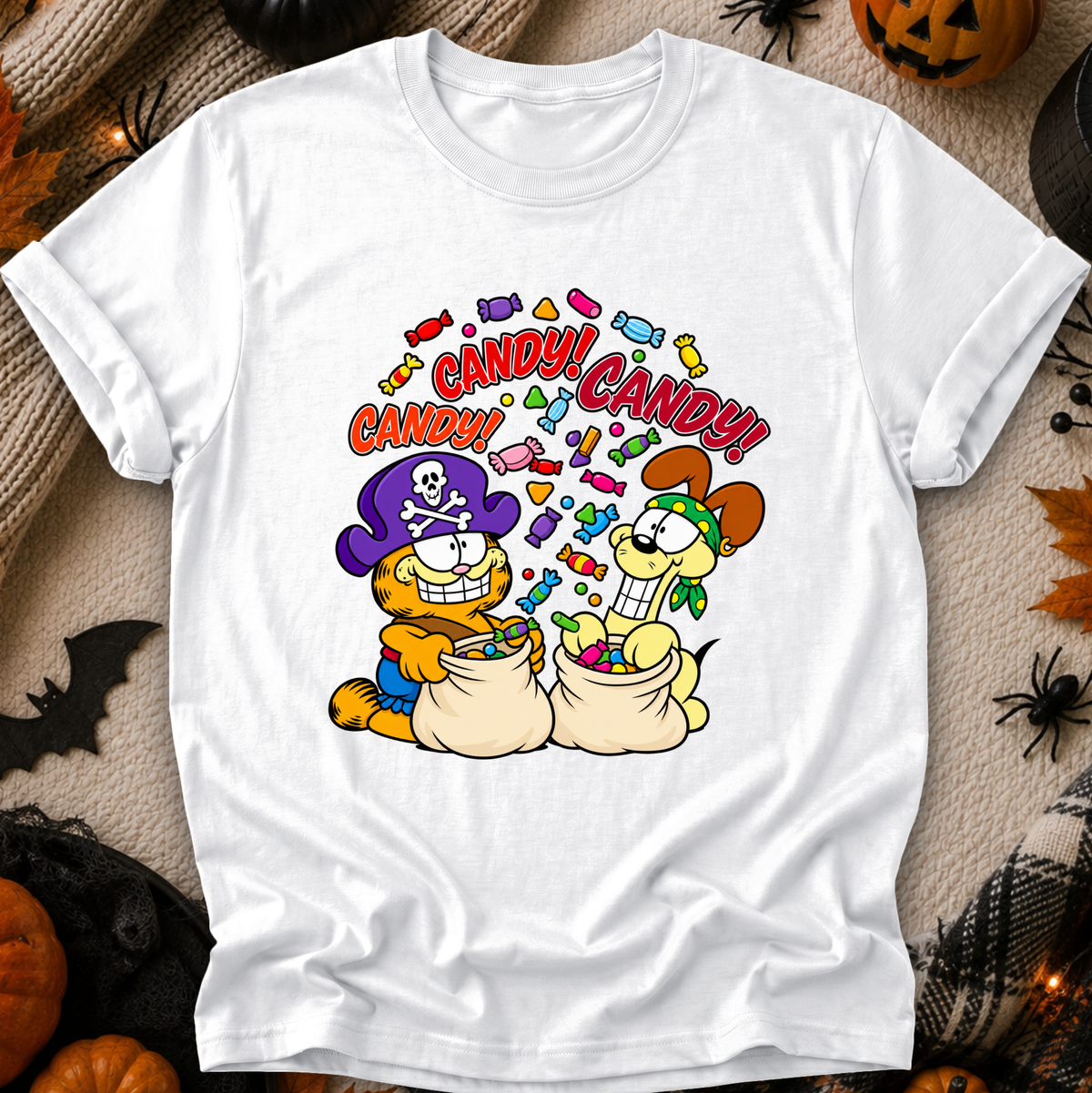 A white short-sleeve crewneck t-shirt featuring a colorful cartoon graphic of Garfield and Odell dressed as pirates with candy bags, surrounded by the text 'CANDY! CANDY!'. The shirt is displayed flat on a cozy, Halloween-themed background with pumpkins, spiderwebs, and a jack-o'-lantern.