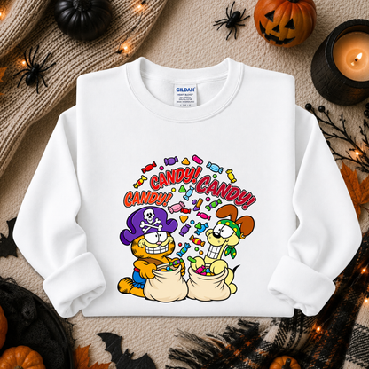 A white long-sleeve crewneck sweatshirt featuring a colorful cartoon graphic of Garfield and Catbus Charlie as pirates with the word 'CANDY' written above them. The sweatshirt is displayed in a flat-lay arrangement surrounded by Halloween decorations including pumpkins, a candle, and a knitted blanket.